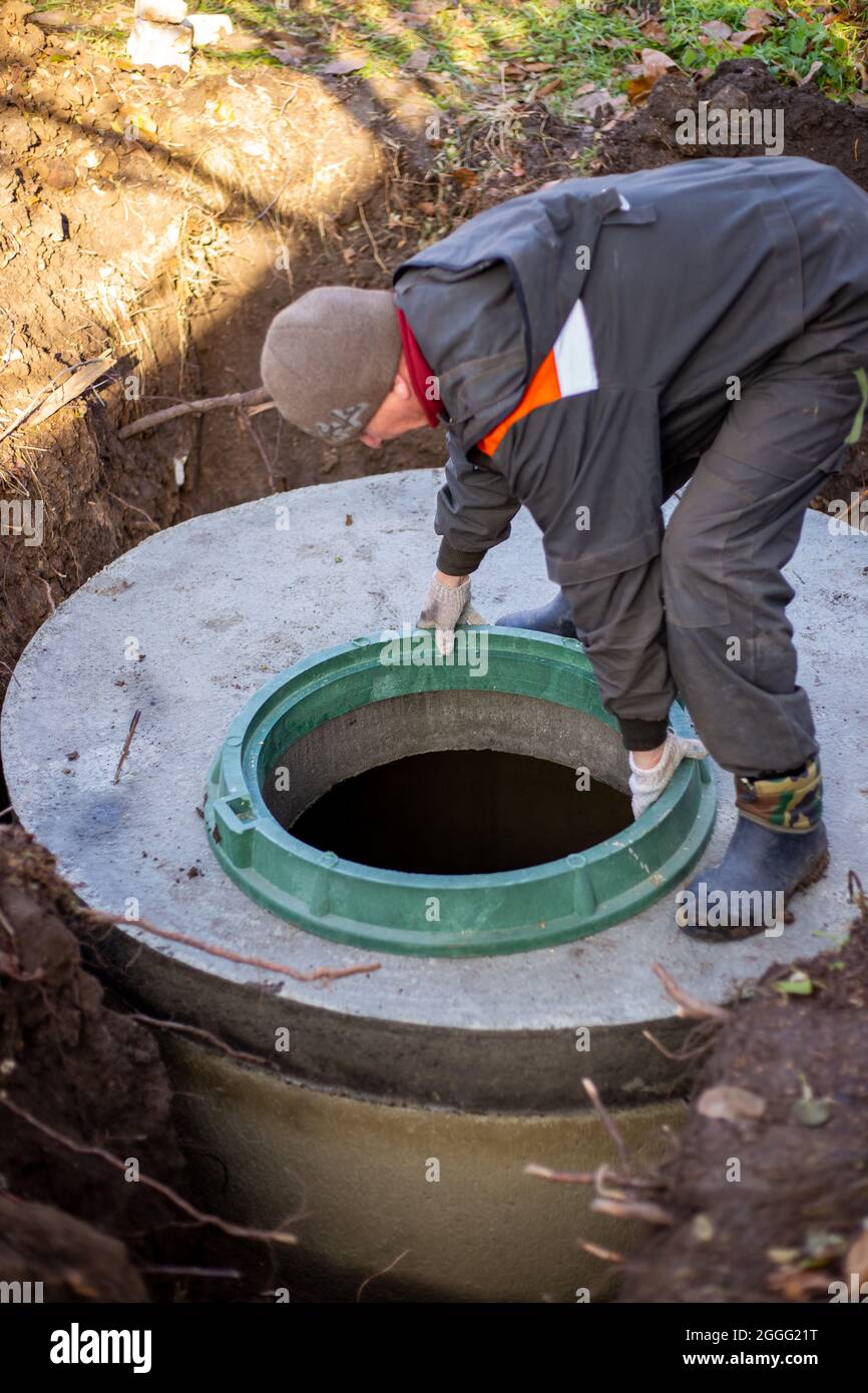 A worker installs a sewer manhole on a septic tank made of concrete rings. Construction of sewage disposal systems for private houses. Stock Photo