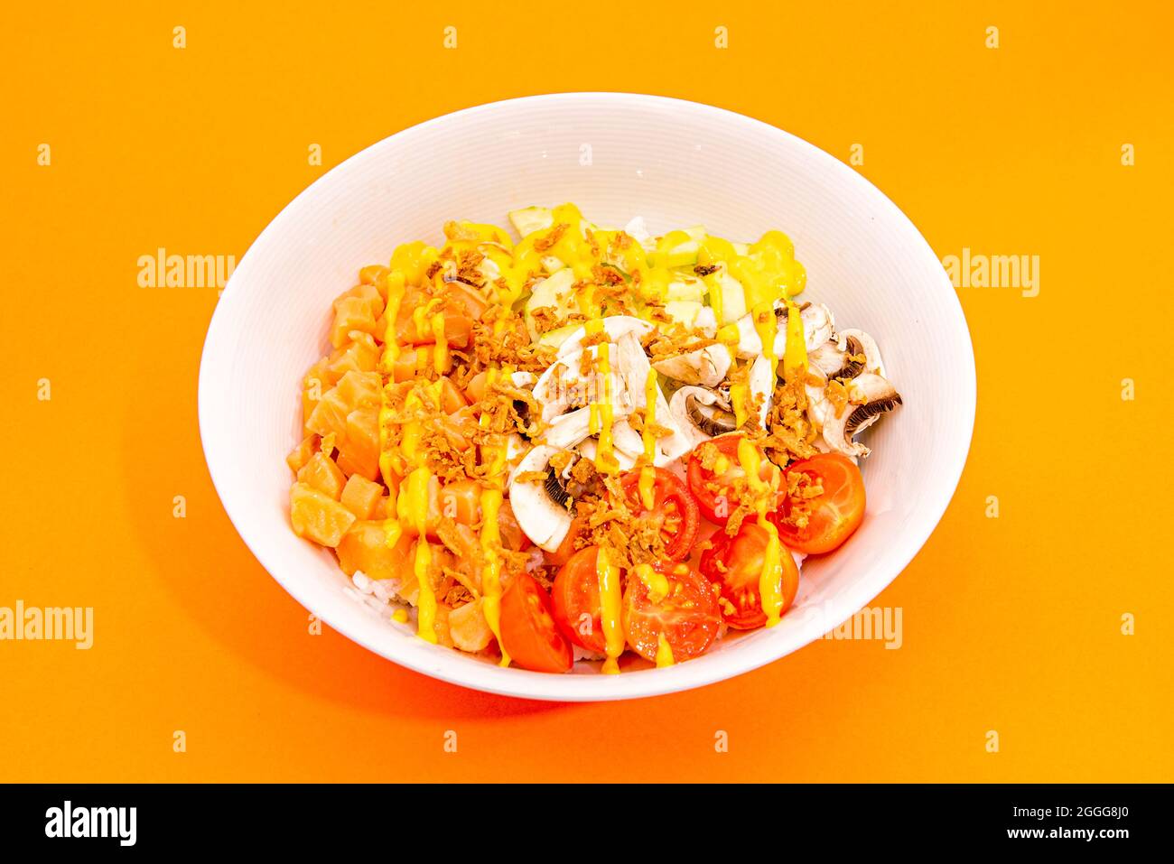 White poke bowl with marinated salmon, pieces of cherry tomatoes, rolled mushrooms, fried onion, cucumber and white rice on orange table Stock Photo