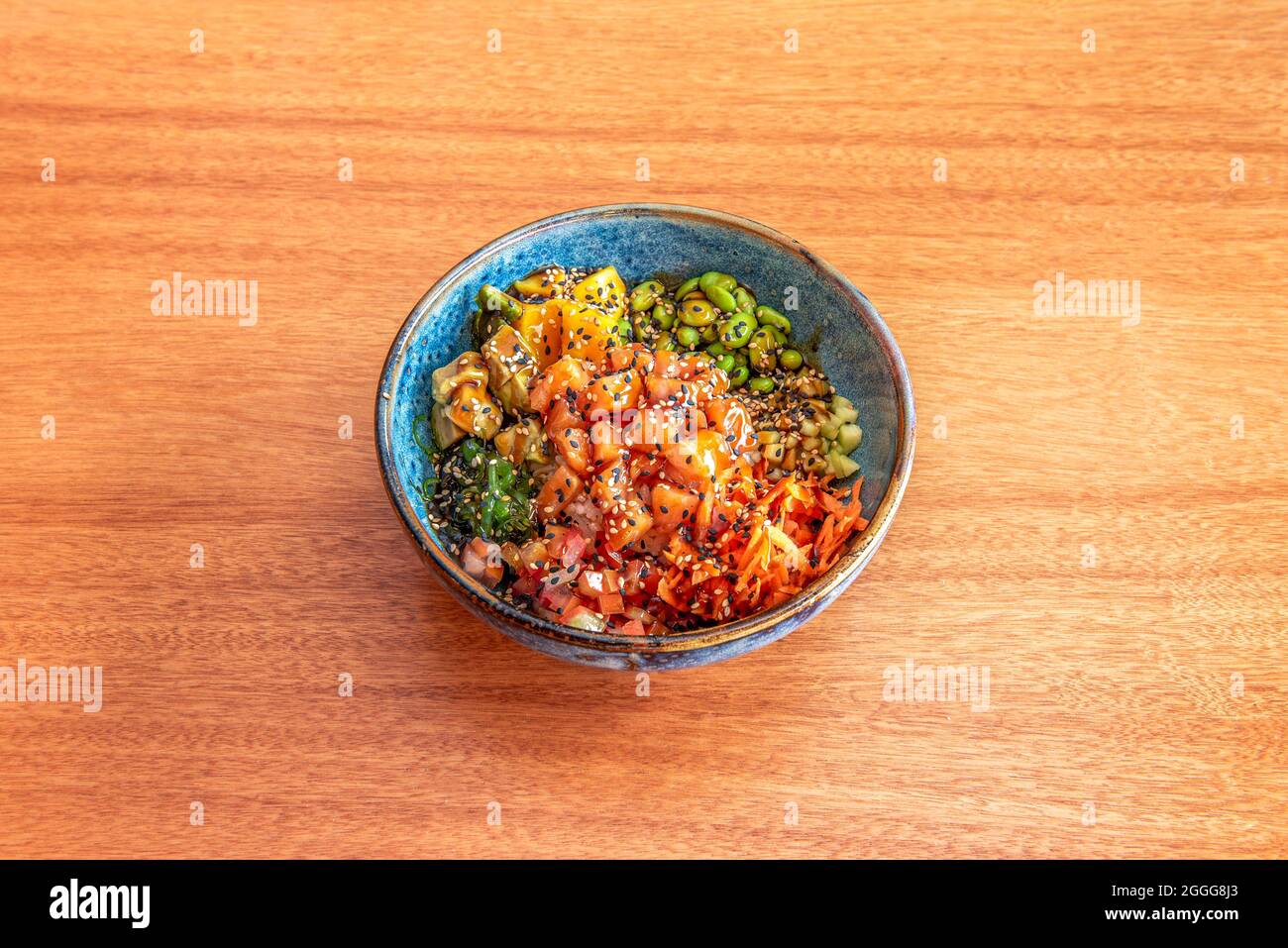 blue bowl of salmon poke with soy sauce, avocado, mango, edamame, wakame seaweed, cucumber and sesame and poppy seeds on cherry wood table Stock Photo