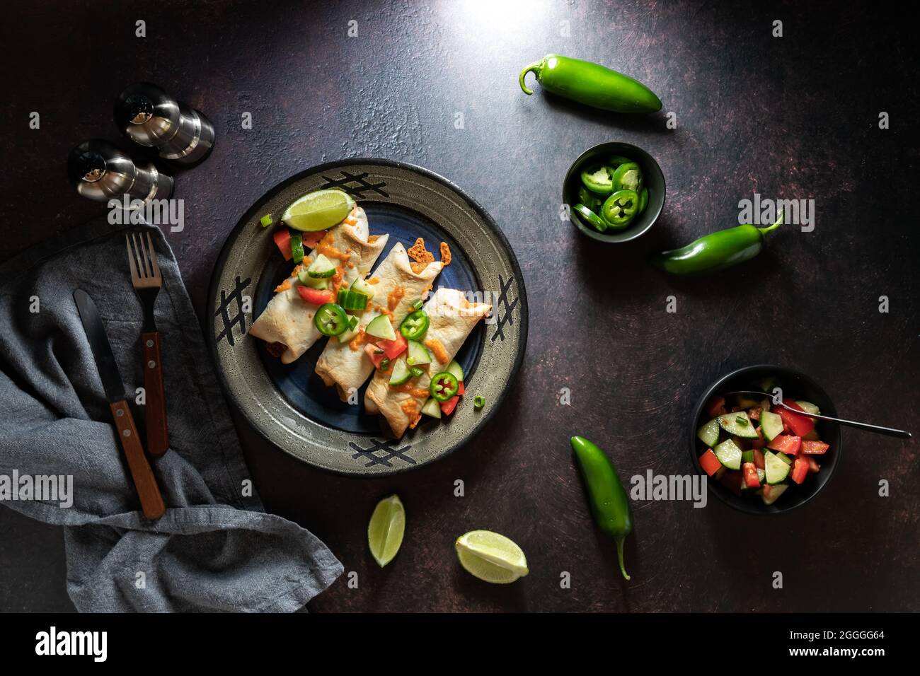 Top down view of a dish of taquitos surrounded by ingredients, against a dark background. Stock Photo