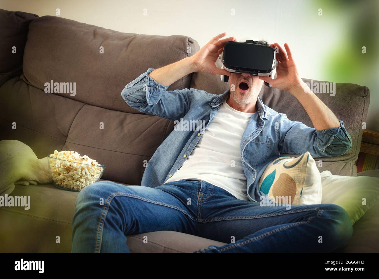Surprised man watching a movie in virtual reality glasses sitting with popcorn on the sofa at home Stock Photo