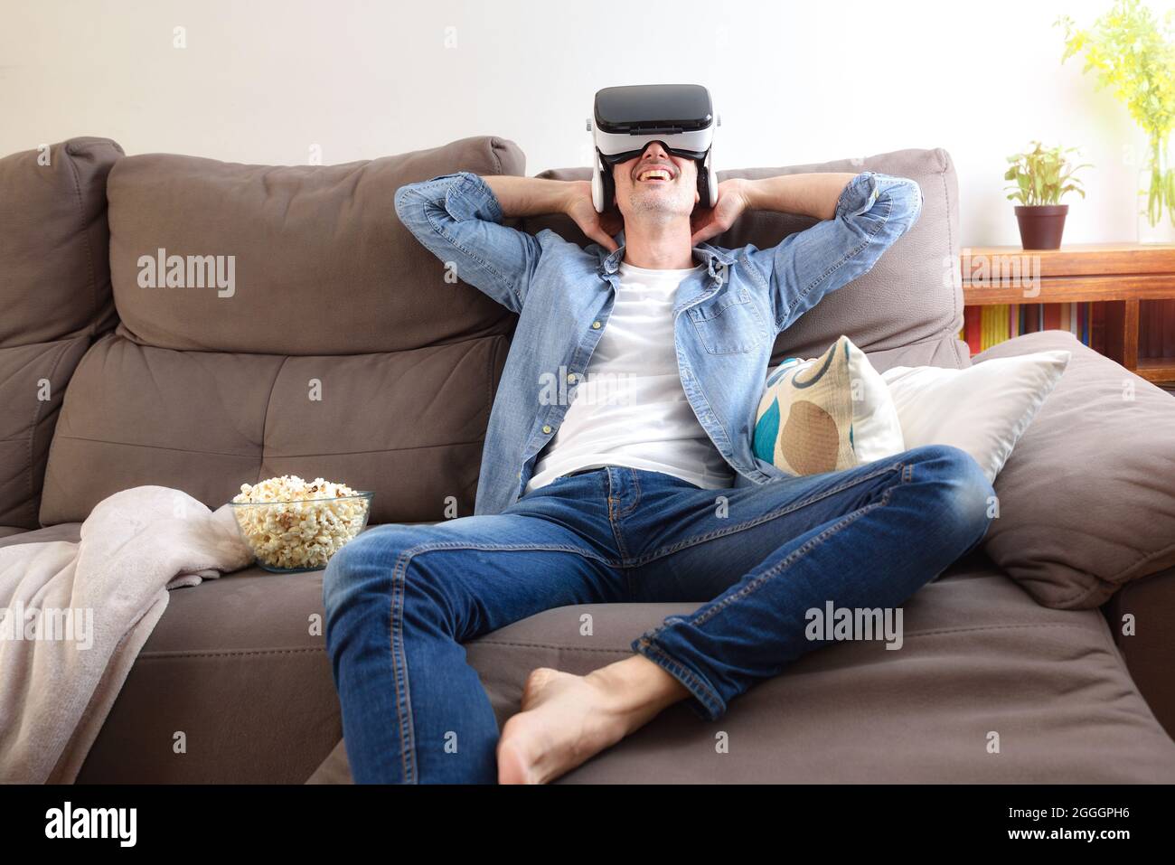 Relaxed entertaining man at home watching movies in virtual reality glasses sitting on the couch with popcorn Stock Photo