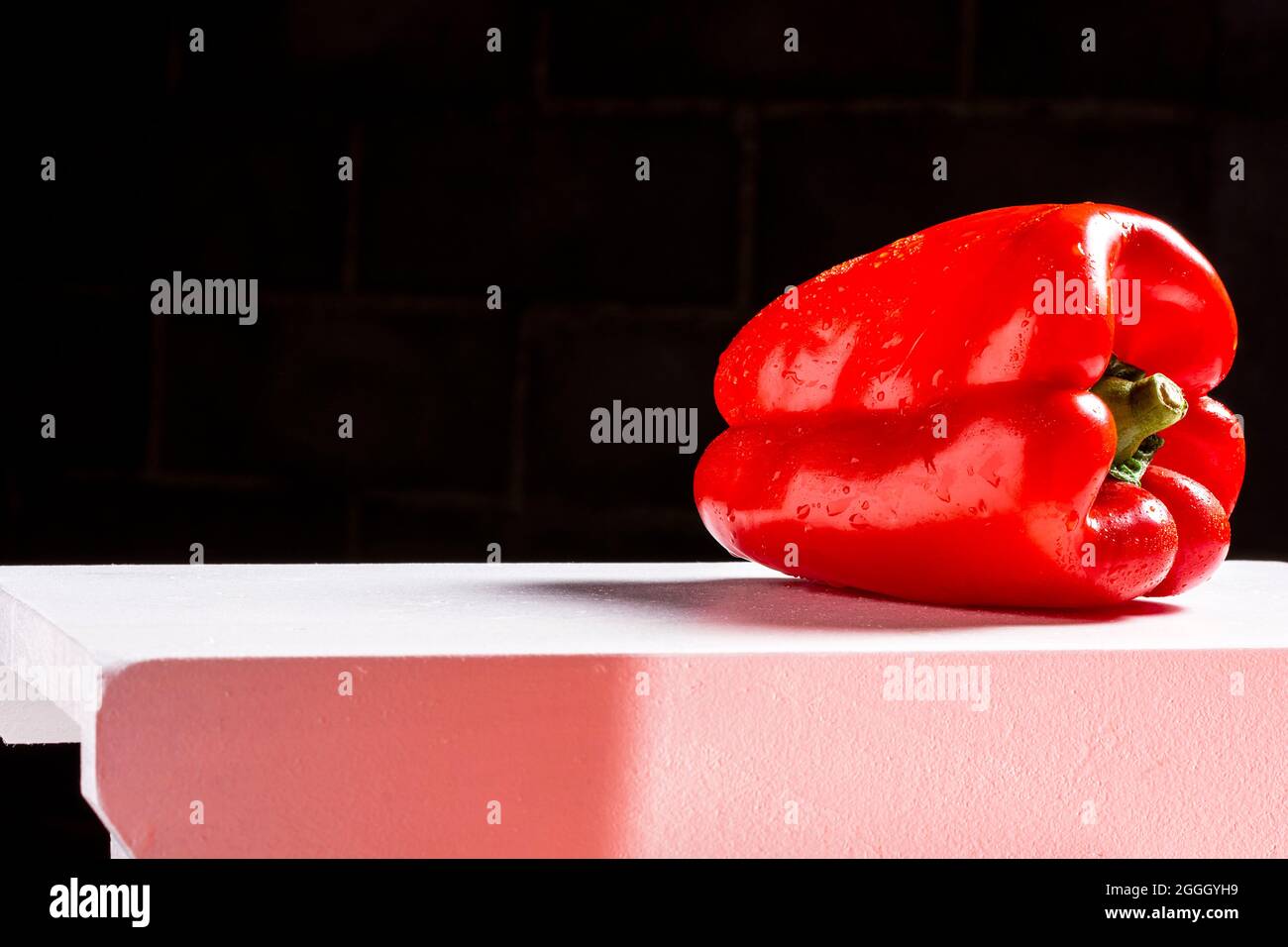 Photograph of a red pepper that is wet and lying on a white bench and a black background.The photo is taken in horizontal format and has space to put Stock Photo