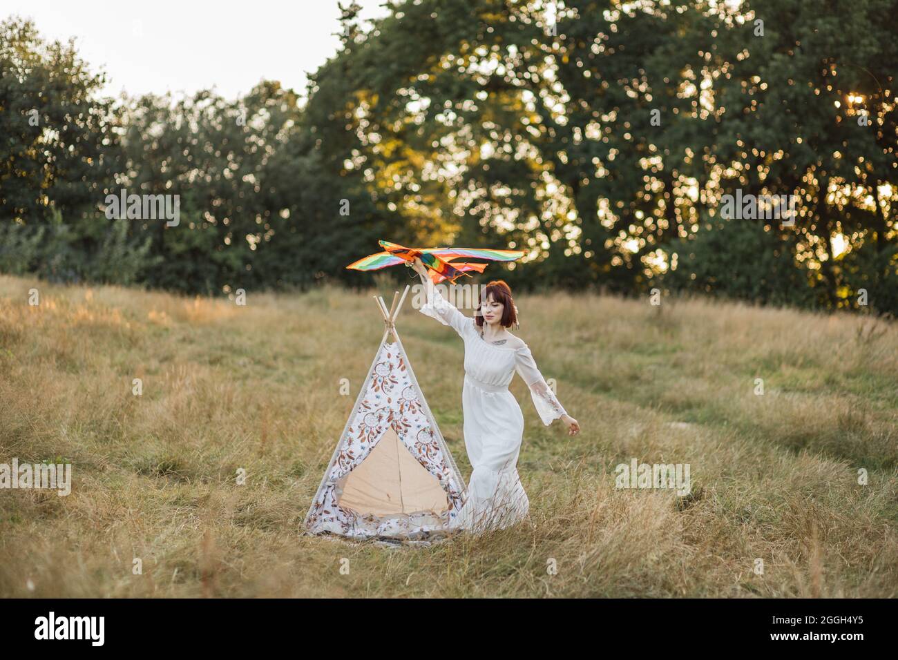 Happy young boho hippie woman running with a kite in the field at sunset in  summer. Stunning indian woman launching colorful kite outdoors in the  meadow. Tee-pee wigwam behind Stock Photo -