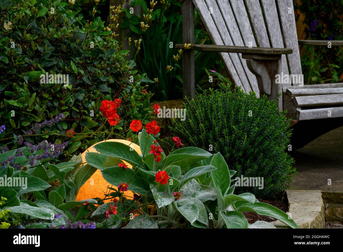 Stylish illuminated globe light lit up & glowing in dark, flowering plants & wooden seat in landscaped private garden at dusk - Yorkshire, England, UK Stock Photo