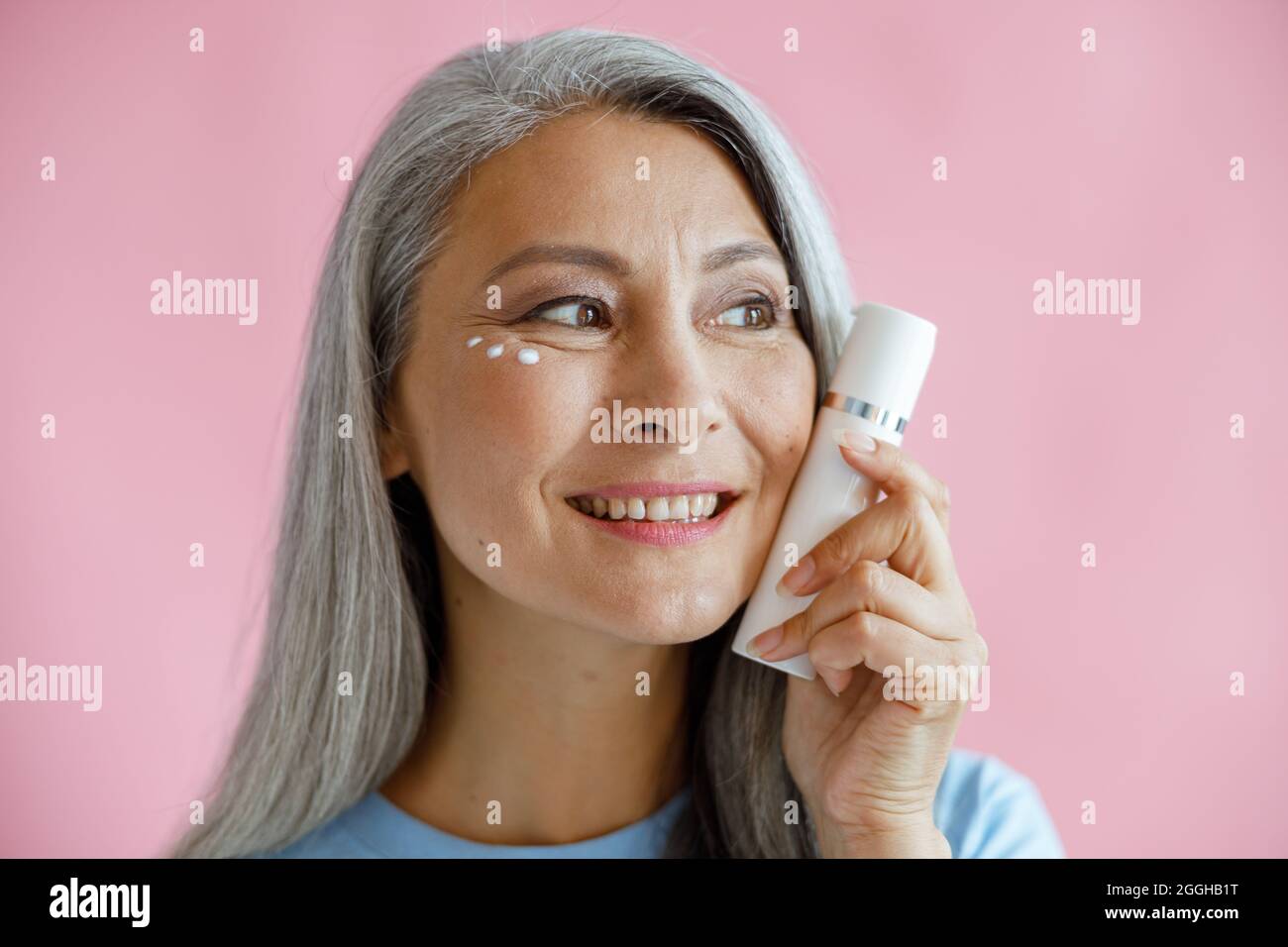 Positive middle aged Asian model with cream drops under eyes holds bottle on pink background Stock Photo