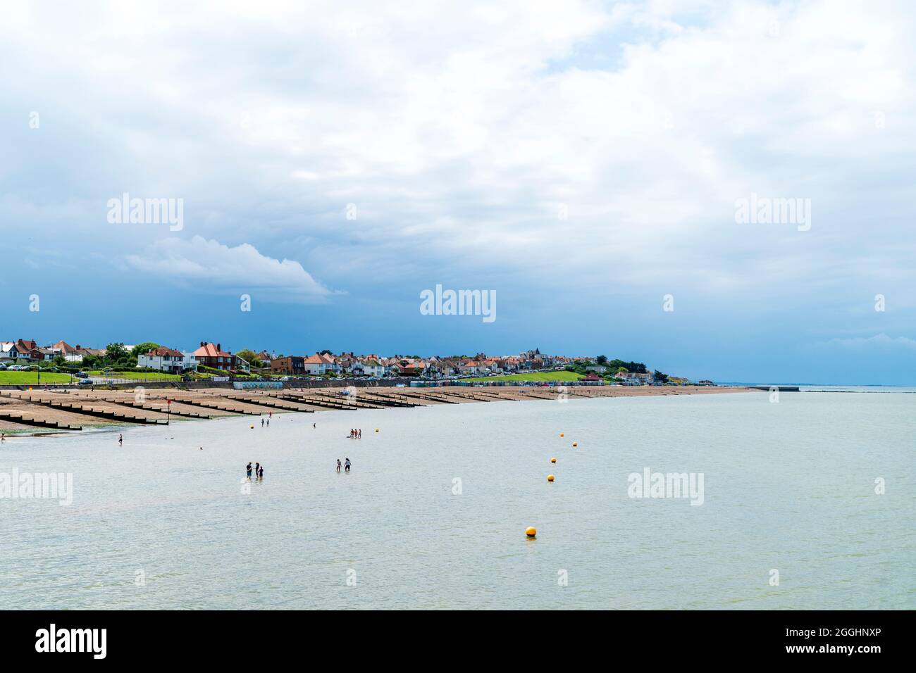 View along the shingle beach with groynes, and the residential part of the town. Dark rain clouds over the town, with white and blue sky over the sea. Stock Photo