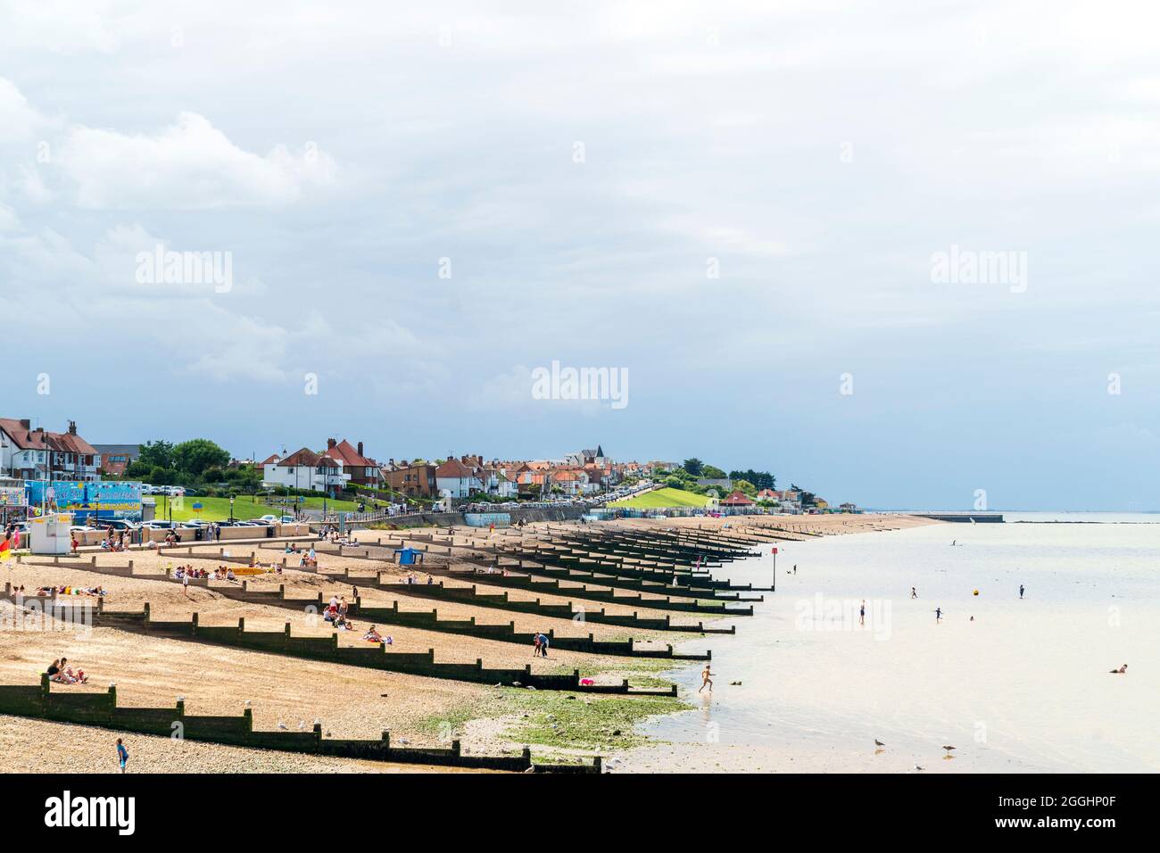 View along the shingle beach with groynes, and the residential part of the town. Dark rain clouds over the town, with white and blue sky over the sea. Stock Photo