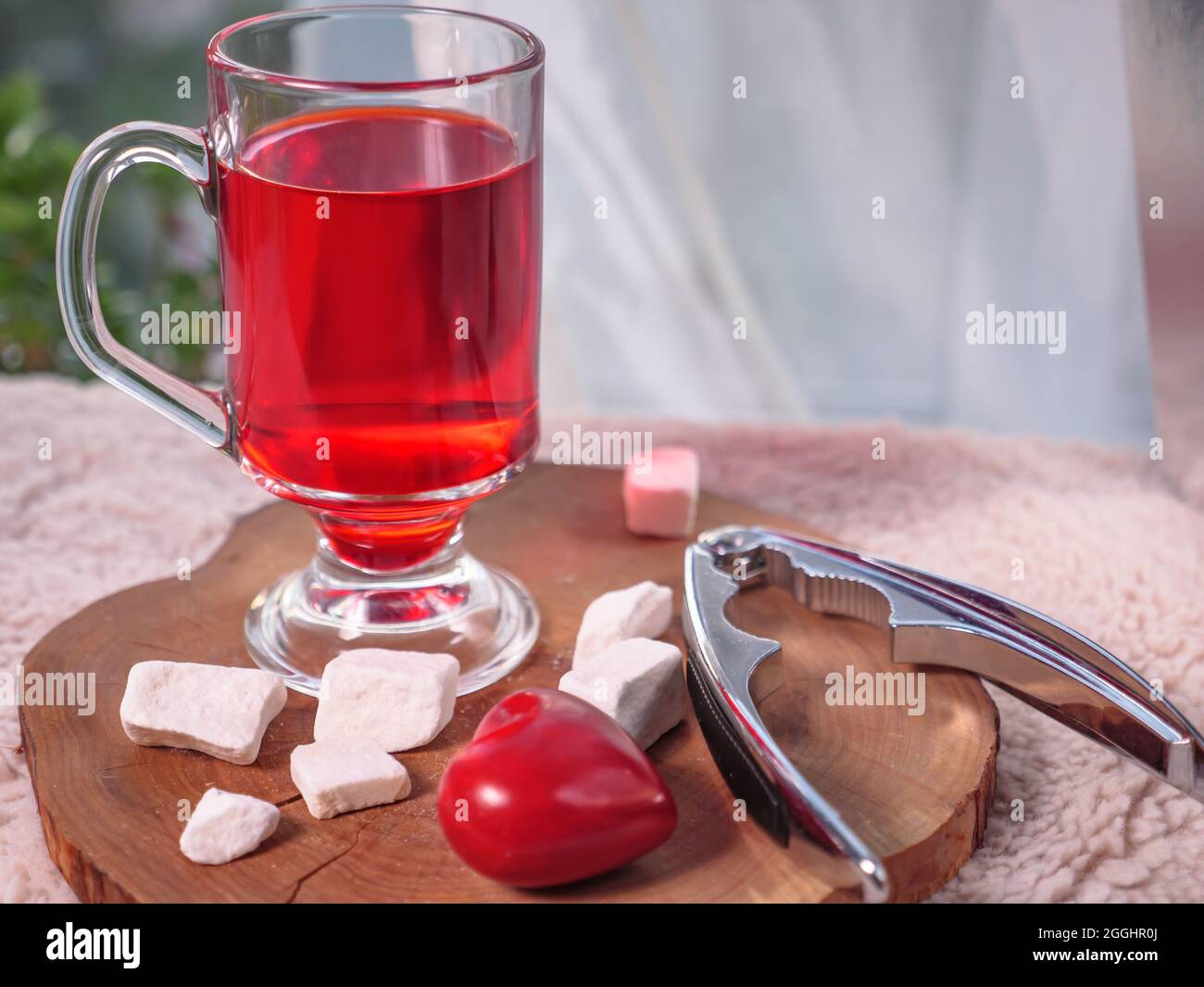 A glass of red berry tea and hazelnuts with nutcracker lying on a wooden board on a cozy orange warm cloth. Window with curtain lace at the background Stock Photo