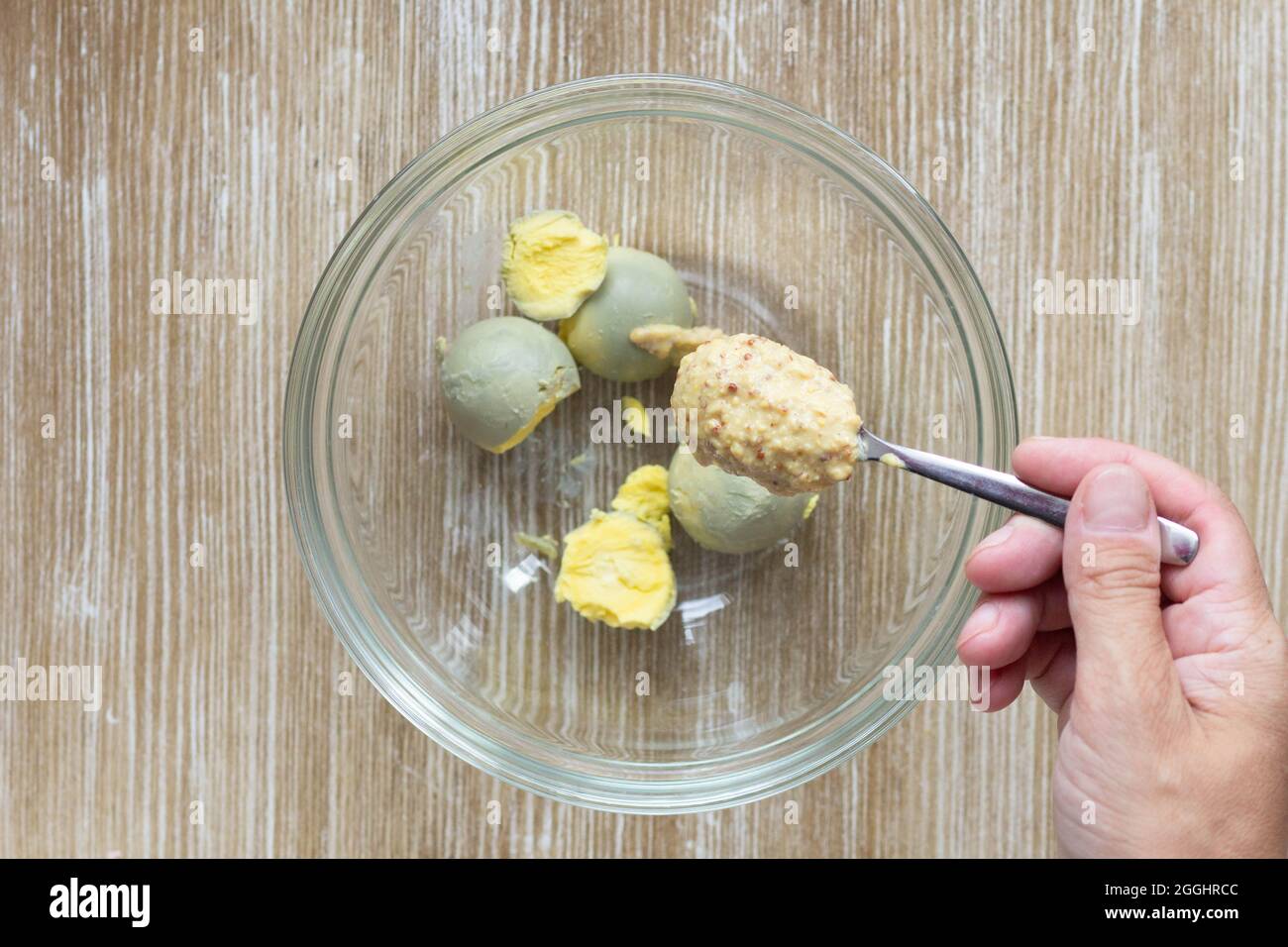 Top view of woman hand holding spoon with mustard to put it into boiled yolks in glass bowl for making flavoring for okroshka Stock Photo