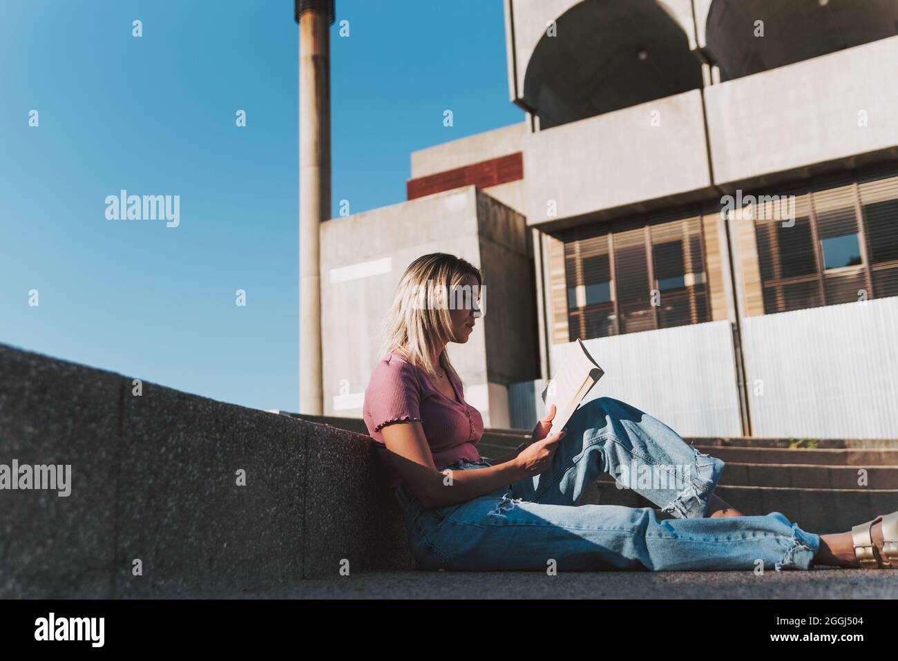 Blonde girl lying down reading a book in an urban environment. Concrete building in the background. Stock Photo