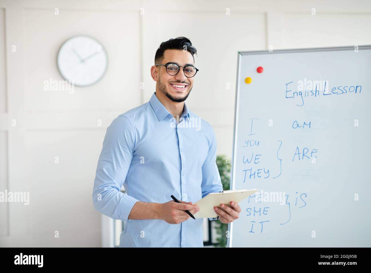 Arab male English teacher explaining rules near blackboard, standing with clipboard, smiling at camera in office Stock Photo