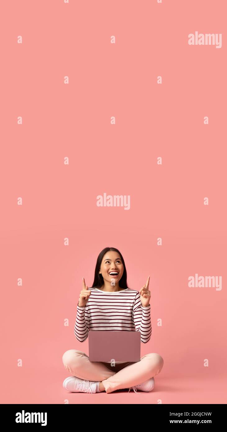 Check This. Excited Asian Female Sitting With laptop And Pointing Up At Copy Space Above Her Head On Pink Studio Background, Cheerful Young Korean Stock Photo