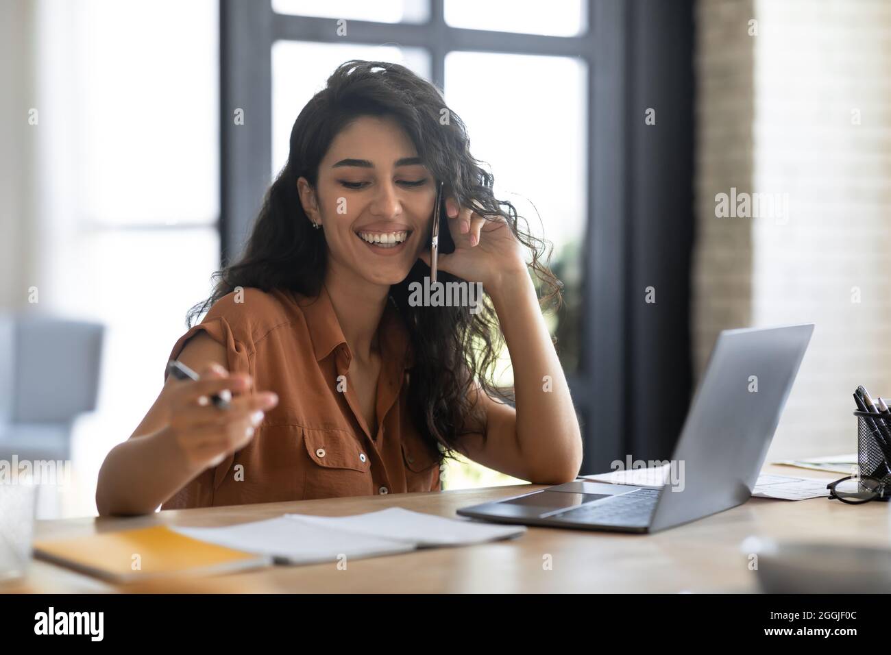 Young business lady talking on cellphone and working on laptop from home office, discussing work issues remotely Stock Photo