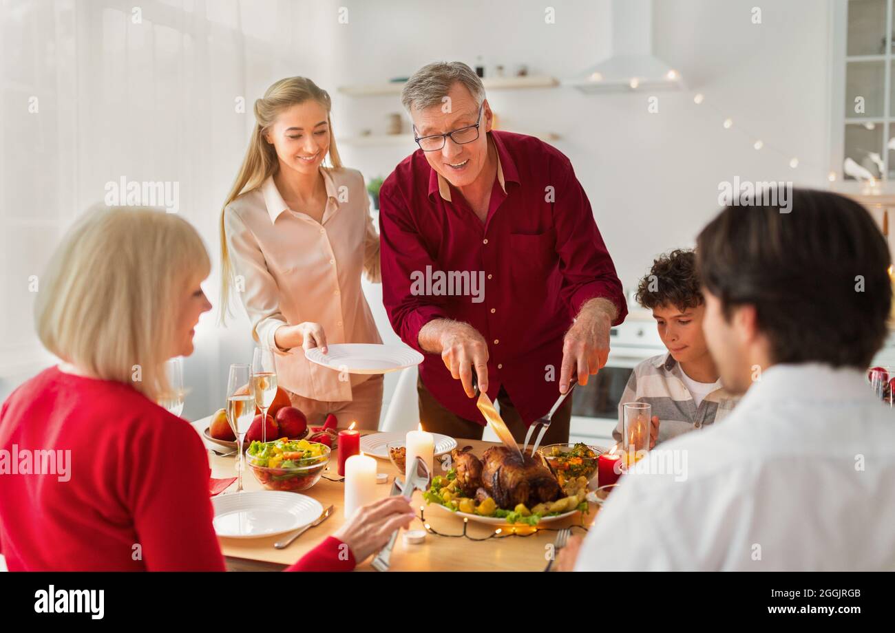 Happy elderly man cutting festive roasted turkey, celebrating Thanksgiving or Christmas with his family at home Stock Photo