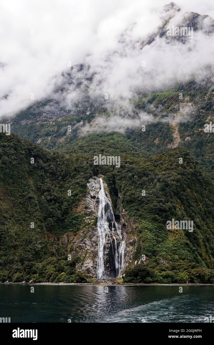 waterfall, moving water, cascading, rock wall, nature, fog above, hillside, green vegetation, yellow kayaks, Milford Sound, Fiordland National Park,Te Stock Photo