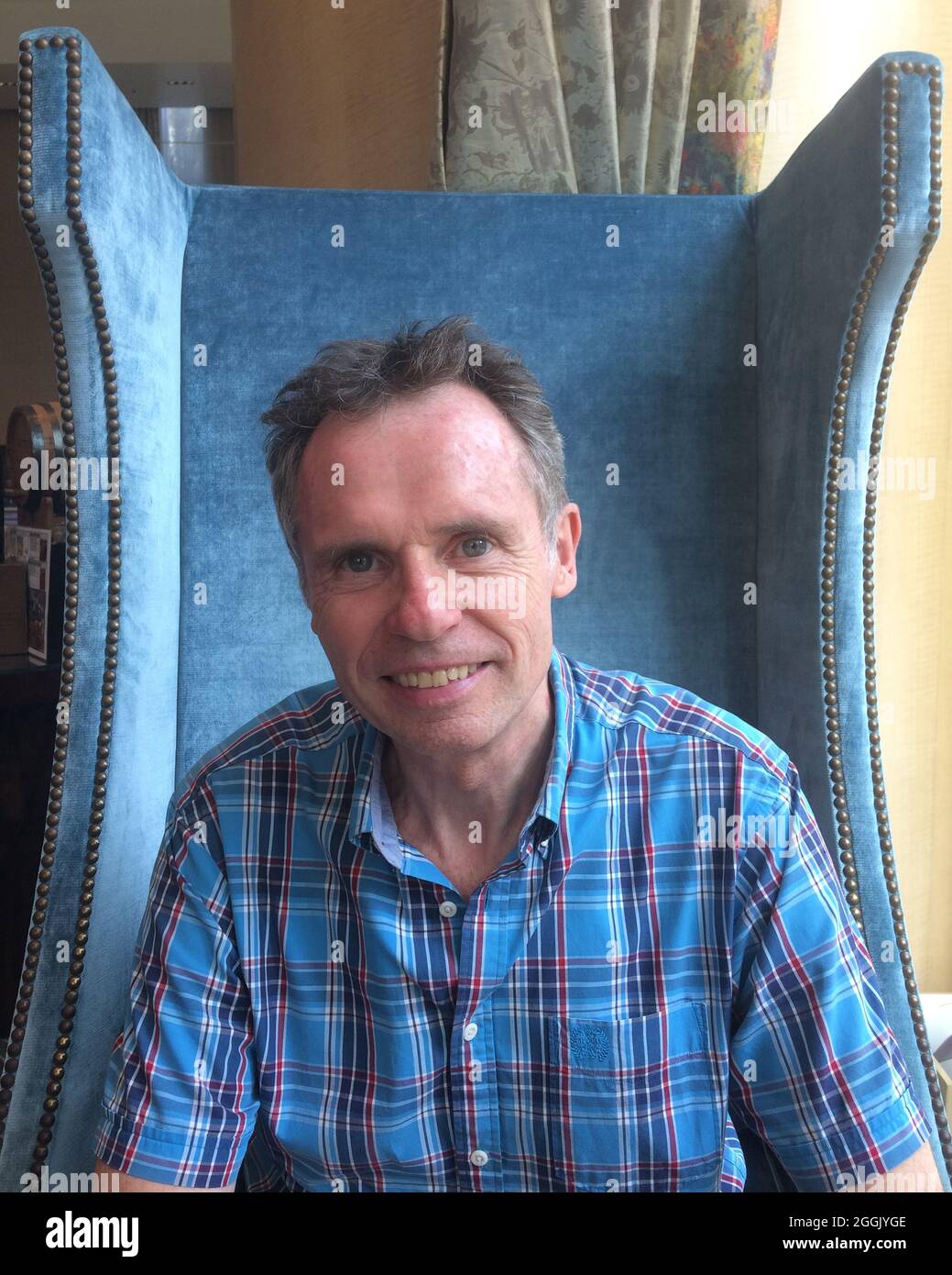 Vertical shot of a Caucasian man sitting on a big blue velvet chair and smiling into the camera Stock Photo