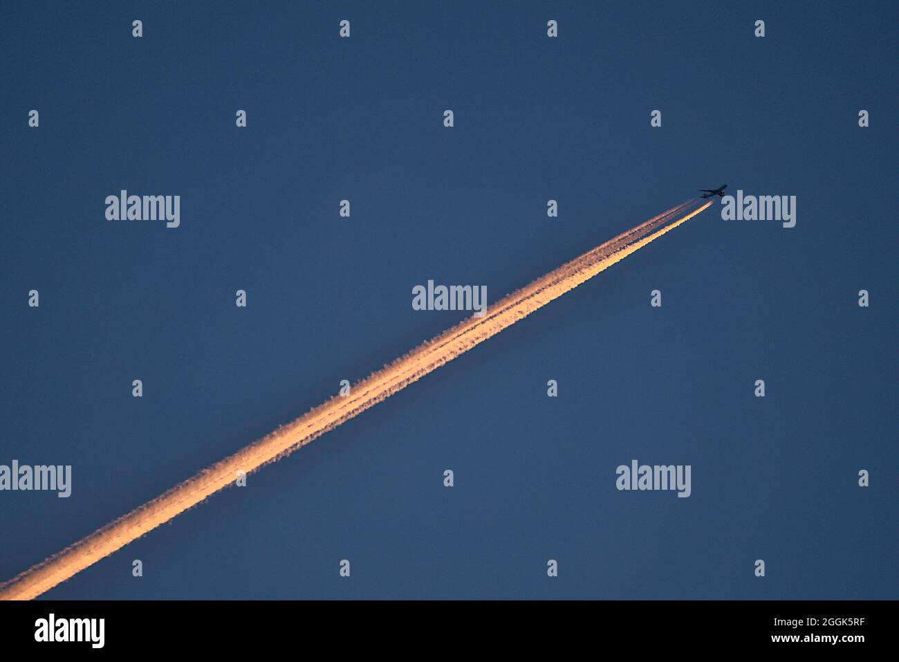 Beautiful telephoto evening view of airplane contrail in orange colors of setting Sun on dark blue background seen over Dublin Mountains, Ireland Stock Photo
