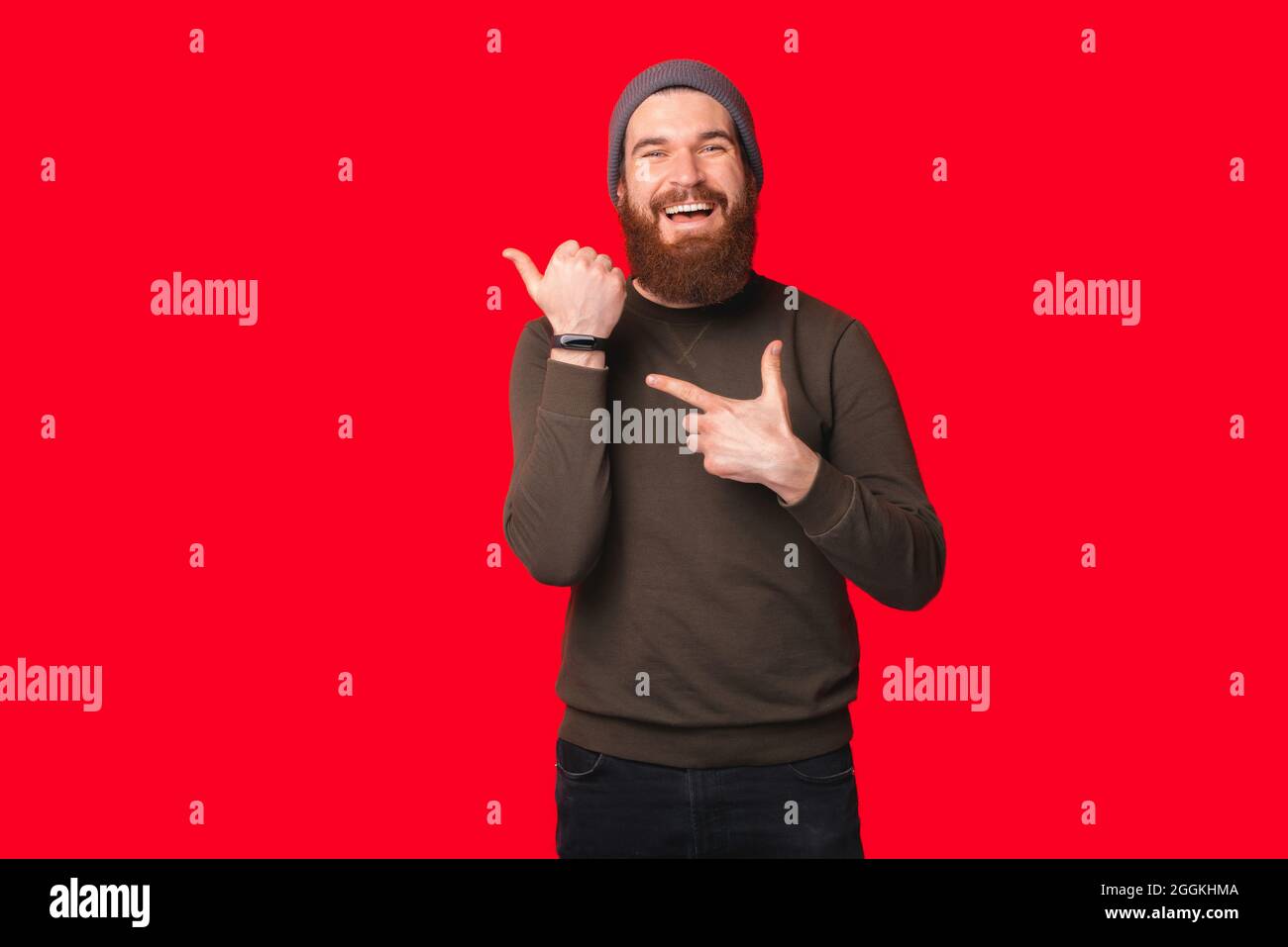 Bearded man wearing warm hat and sweater is pointing at his wrist watch over red background. Stock Photo