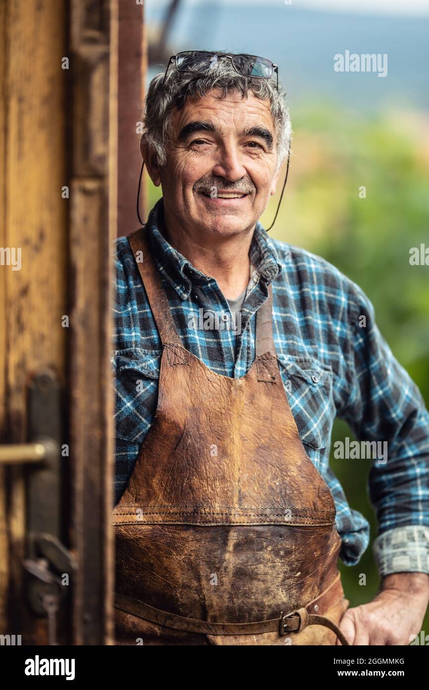 Older and experienced craftsman stands smiling in front of the workshop in checkered shirt and leather apron. Stock Photo