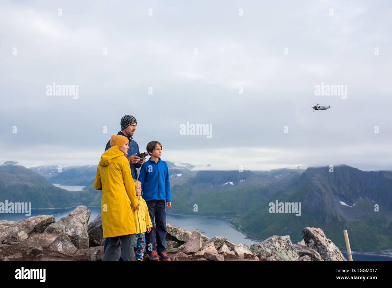 Happy family, standing on a rock and looking over Segla mountain on Senja island, North Norway, taking pics with drone. Amazing beautiful landscape an Stock Photo