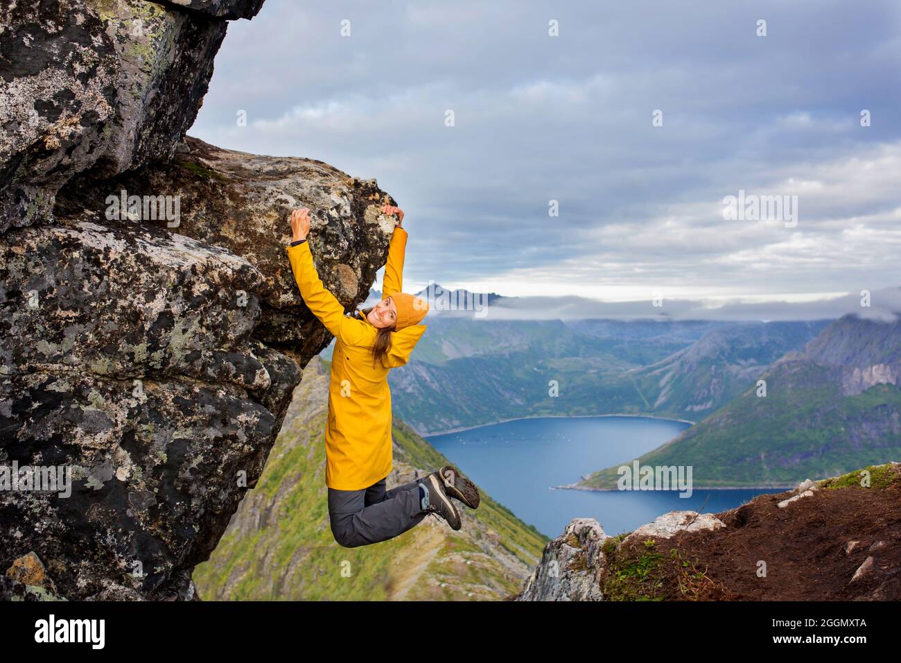 Woman in yellow raincoat, hanging from a rock over Segla mountain on Senja island, North Norway. Amazing beautiful landscape and splendid nature in sc Stock Photo