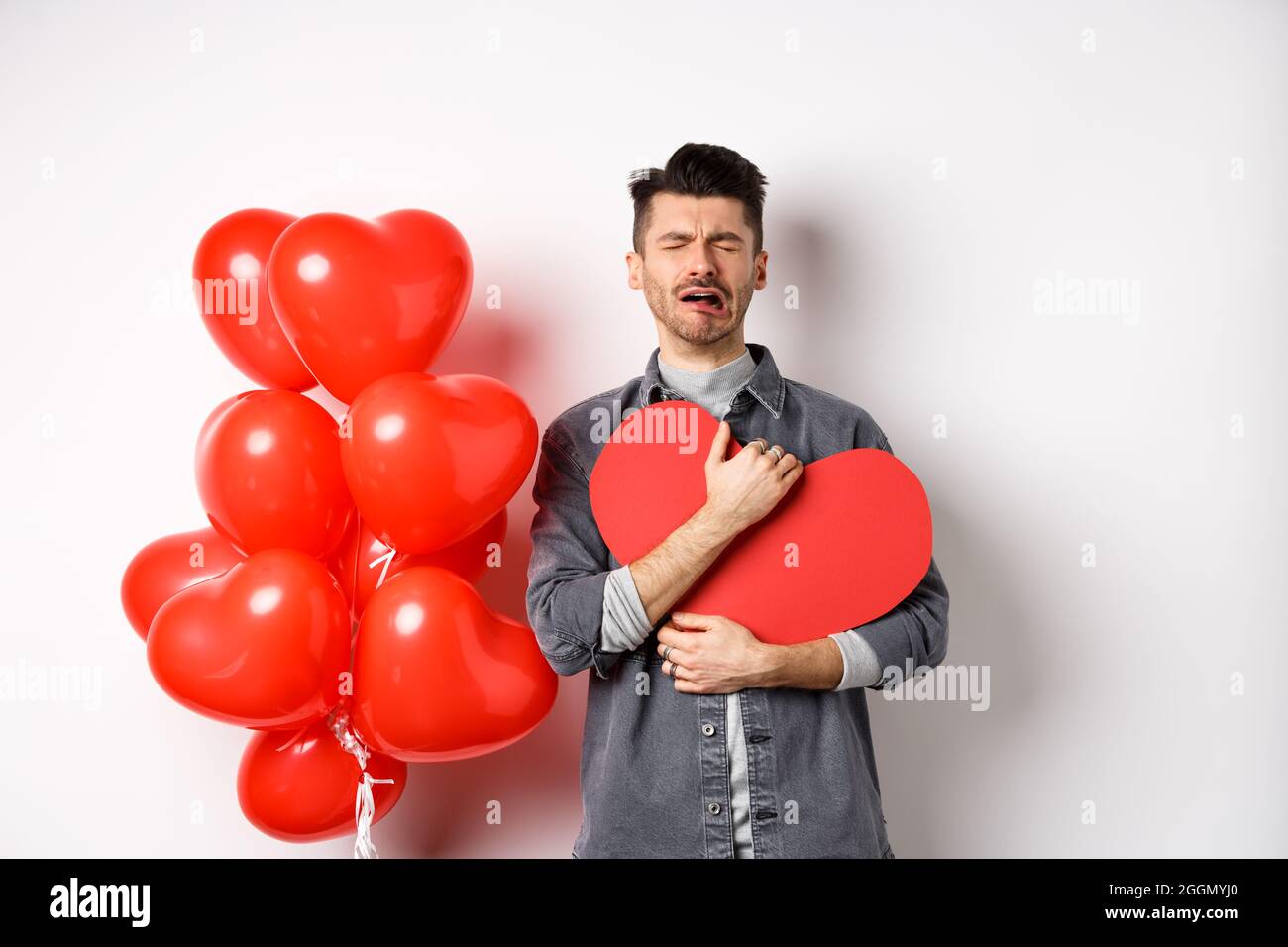Crying man standing single and lonely on Valentines day, hugging heart cutout and sobbing miserable, being heartbroken and rejected by lover, white Stock Photo