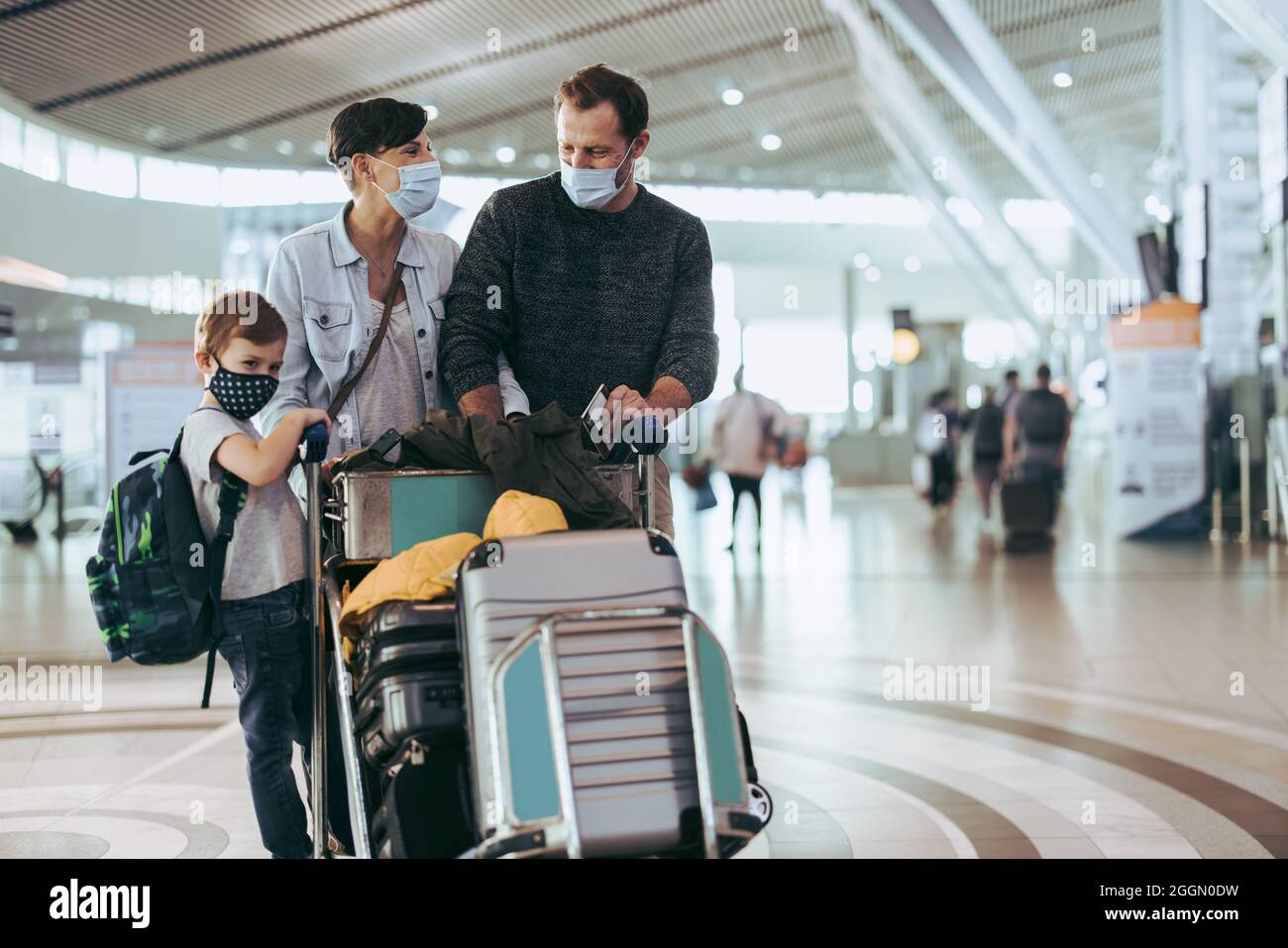 Traveler family wearing face mask with luggage trolley waiting at airport. Couple walking with their son at airport in pandemic. Stock Photo
