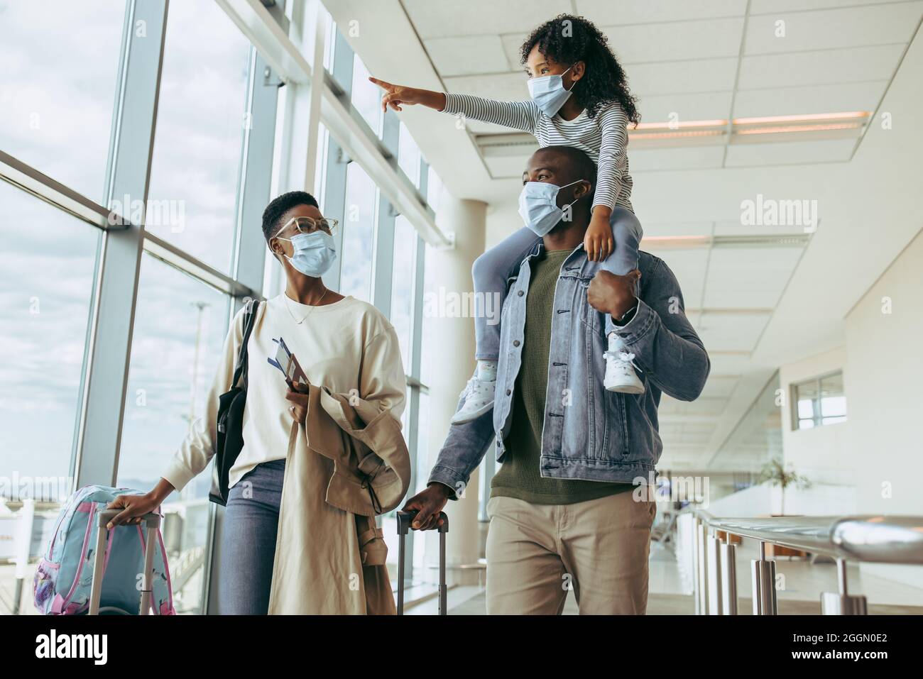 Young girl pointing away while on shoulder of her father and woman at airport. African family traveling during pandemic. Stock Photo