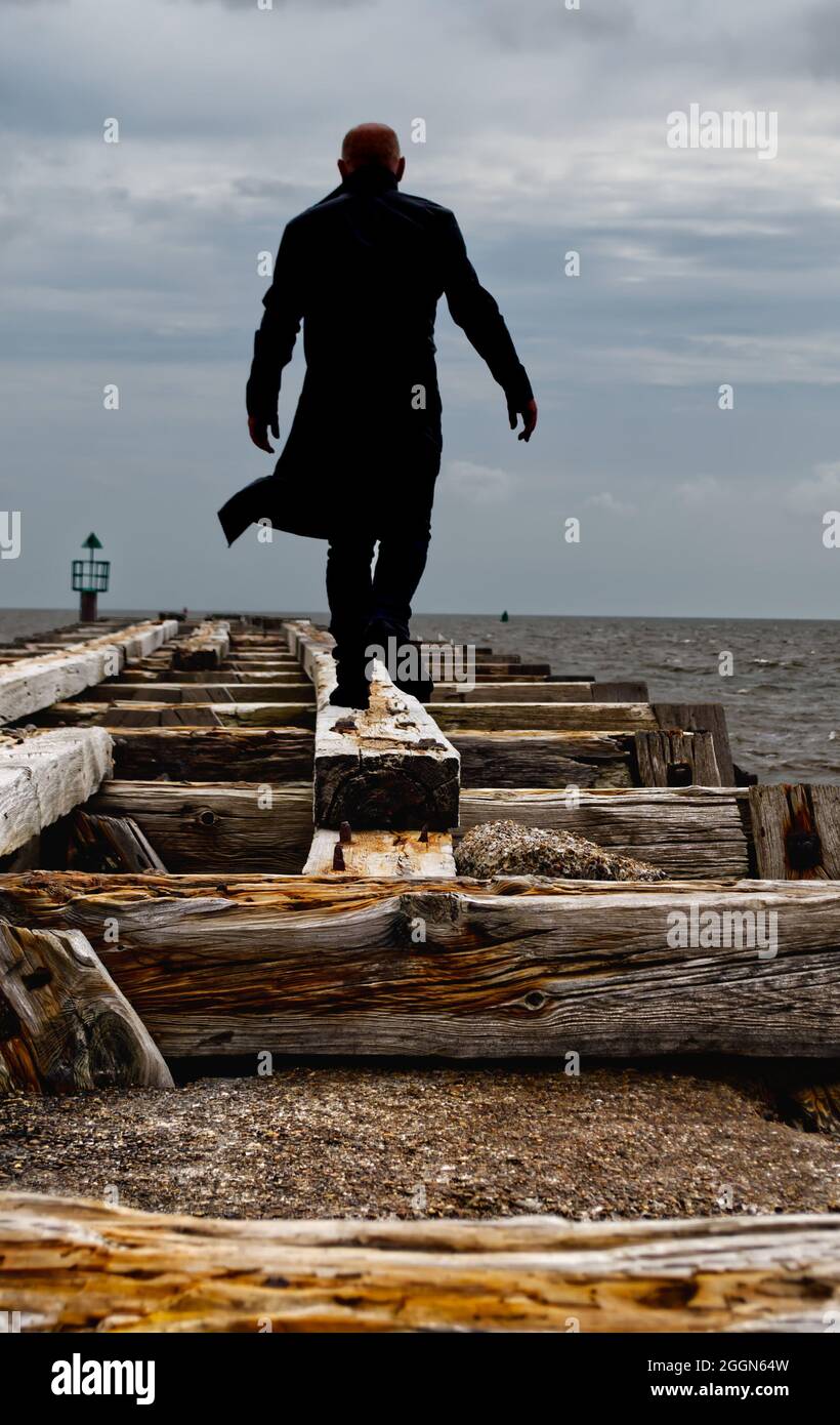 shadowy man in long black coat (mid distance) walking on old beam of ruined pier going towards the sea Stock Photo