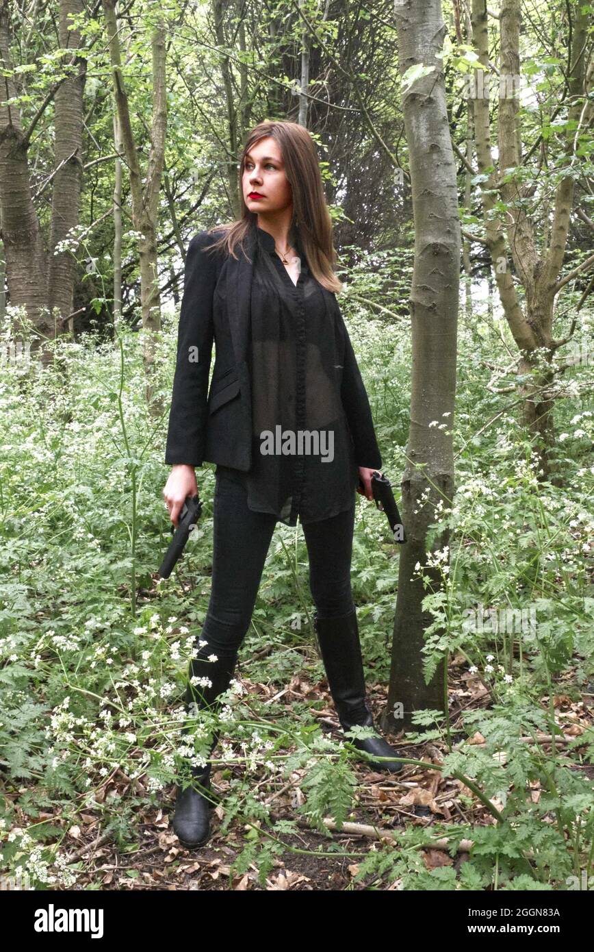 gun toting young woman in the woods dressed in black, female secret agent style. Stock Photo