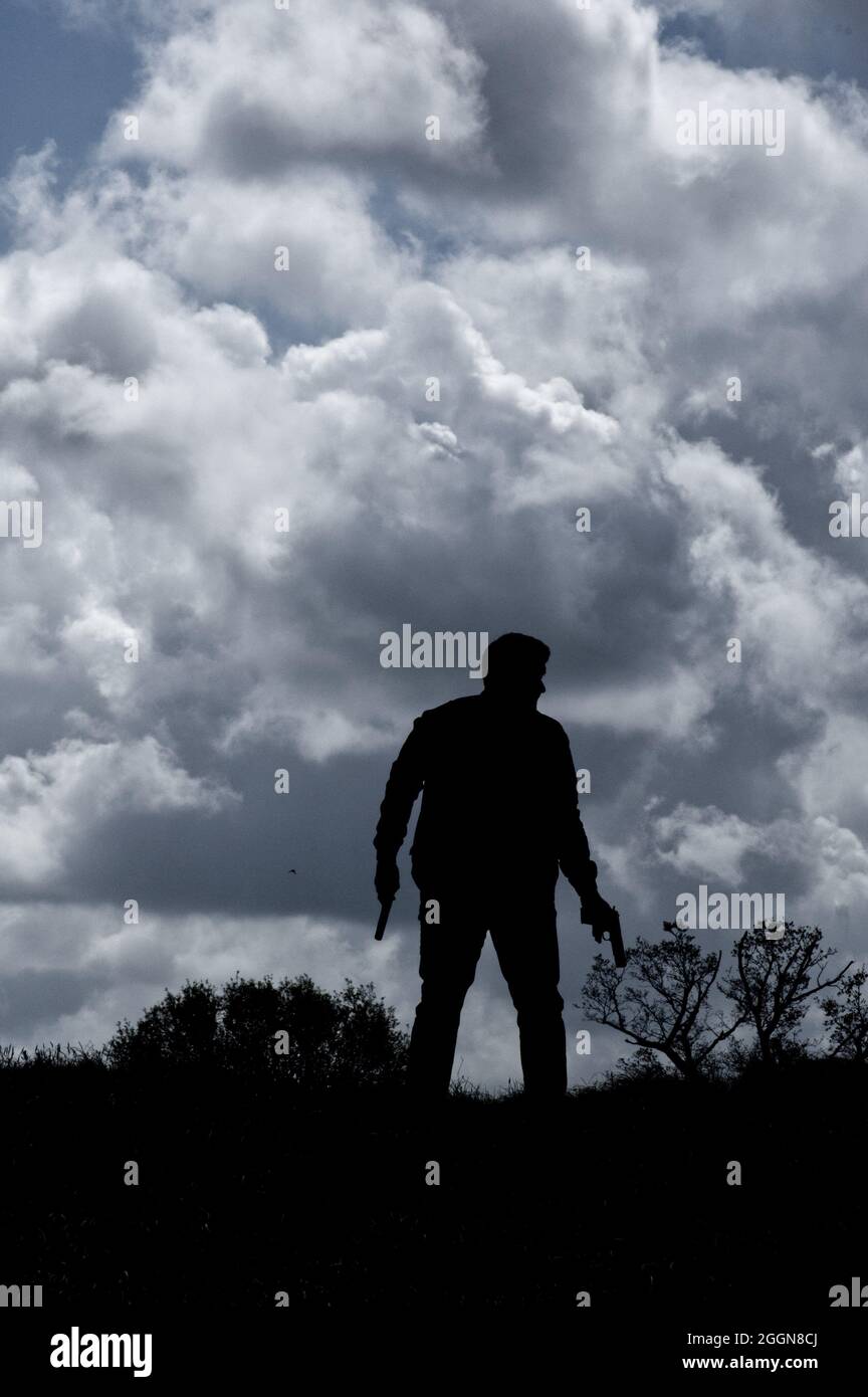 Silhouette of man in country side standing against a cloudy sky, holding a pistol. (book cover style) Stock Photo