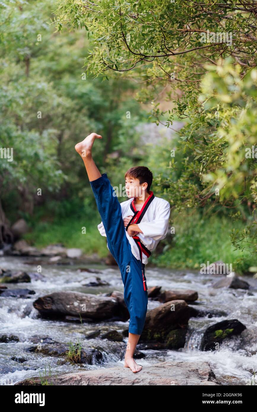 The Japanese child practices taekwondo with a martial arts kimono in nature. Black karate belt training with his leg up. Stock Photo