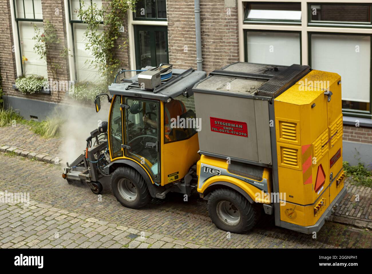 ZUTPHEN, NETHERLANDS - Aug 20, 2021: Steam coming out of weed and tares pavement clean sweeper machine in 19th century cobble stone street. Stock Photo