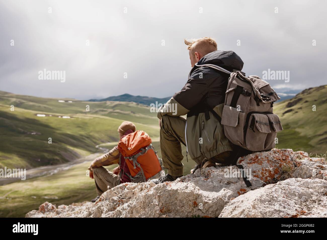 Two guys hikers sits and relax in mountains and looks on green valley Stock Photo