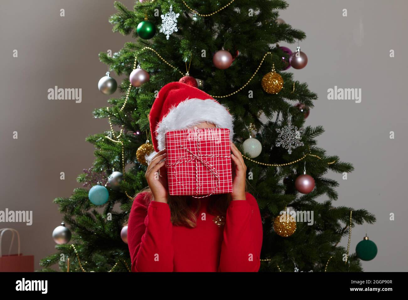 A funny child in a Santa Claus hat hides his face behind a box with a Christmas present. Little girl on the background of a Christmas tree. Stock Photo
