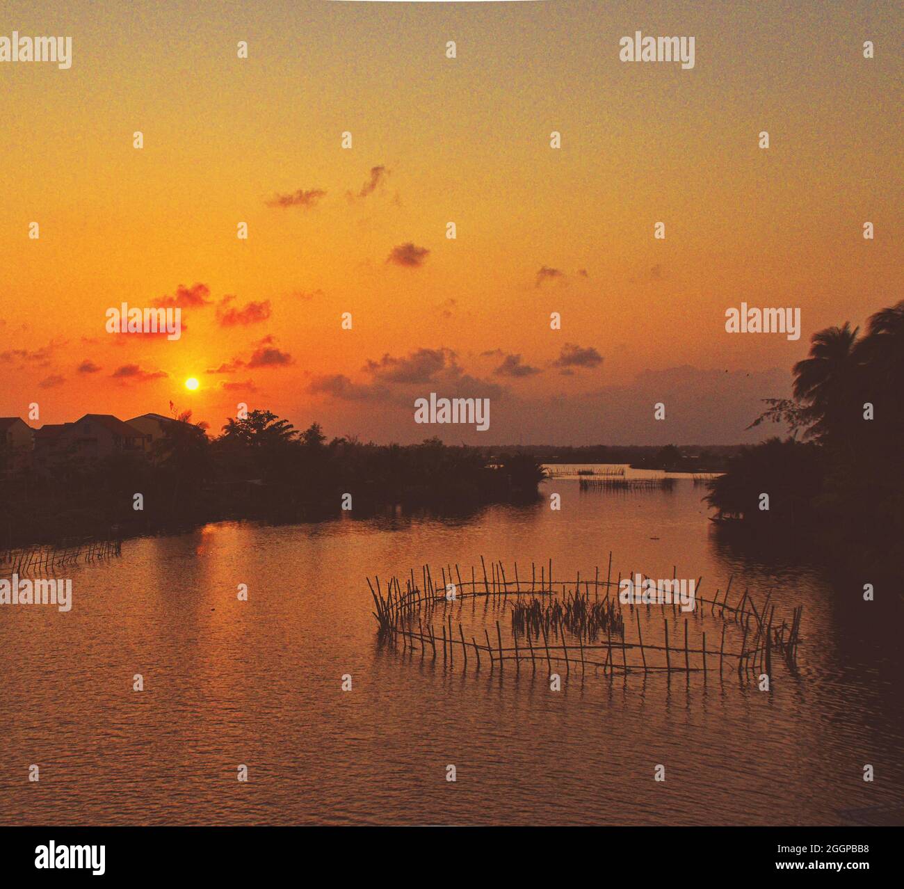 Scenic sunset over a calm river with traditional fishing nets and silhouetted trees, creating a tranquil landscape bathed in warm orange and golden. Stock Photo