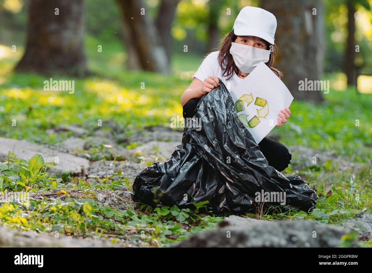 Close up photo of a young girl in a white cap wearing medical face mask  holding a paper with the recycled sign drown on it and gathering into the  Stock Photo -