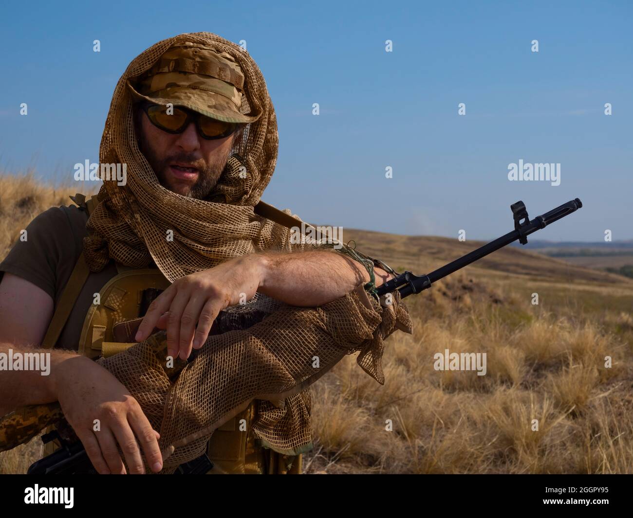 Close-up photo of a mercenary sniper in camouflage clothes under the scorching sun. He stands with a rifle and looking to camera. Concept of a profess Stock Photo