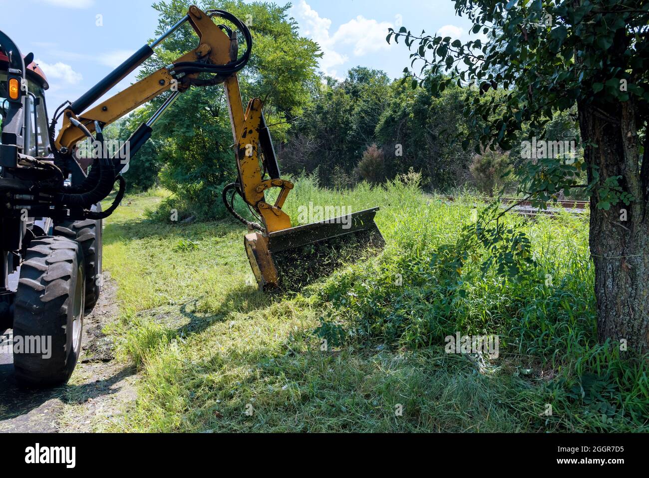 Road services are engaged in tractor with a mechanical mower mowing grass on the side of the asphalt road Stock Photo