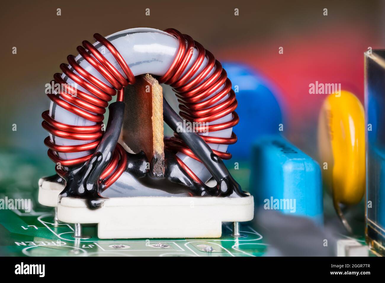 Ferrite core inductor on white holder and colored capacitors on green circuit board. Close-up of electronic toroidal coil with insulated copper wire. Stock Photo