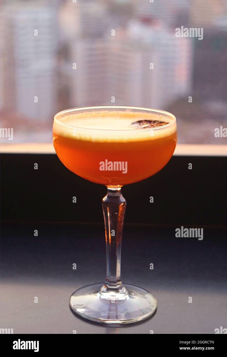 Closeup a glass of vibrant orange cocktail at the rooftop terrace with blurry urban view in background Stock Photo