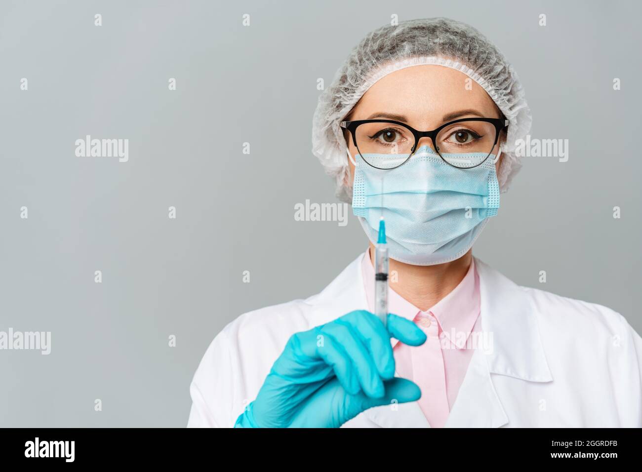 Female doctor or scientist in white medical gown, blue gloves, green cap and mask holds a syringe in hands on white background. She is ready to give Stock Photo