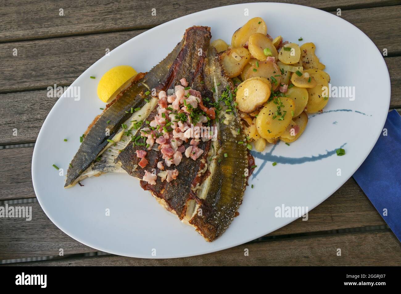 Freshly fried whole plaice from the Baltic Sea with bacon, fried potatoes and chives garnish on a plate on a wooden table, high angle view from above Stock Photo