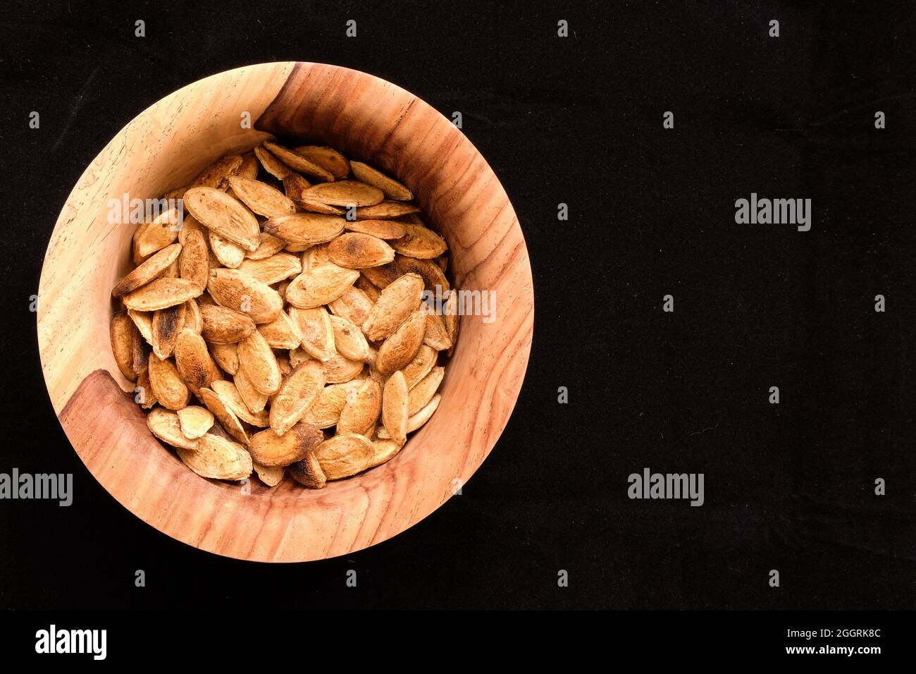Homemade dry roasted unshelled pumpkin seeds with salt in a wooden bowl on a black background; charred pepitas ready for peeling and eating. Stock Photo