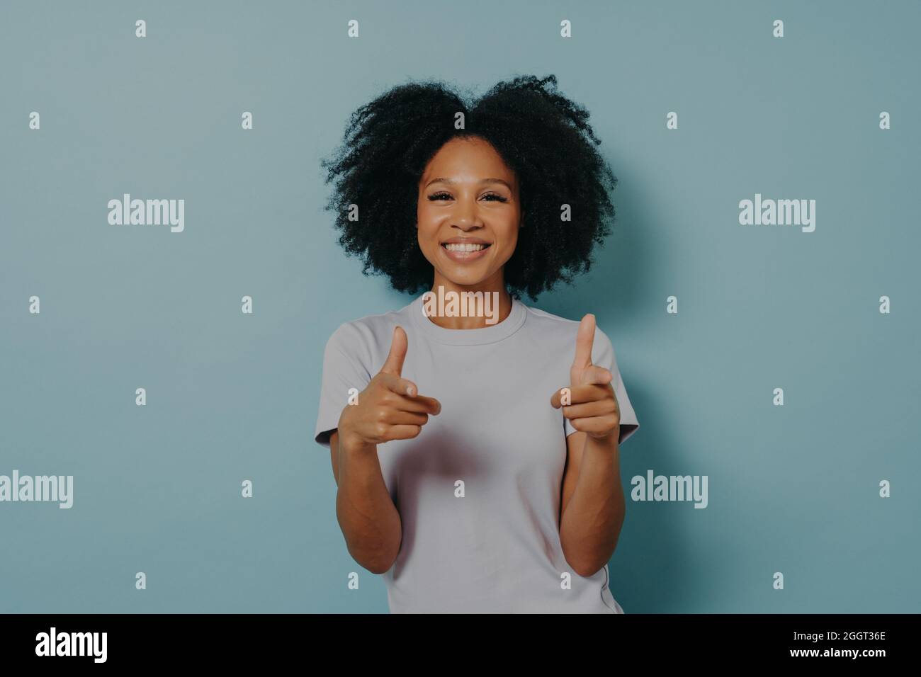 Positive friendly dark skinned lady making finger gun gesture while standing on blue background Stock Photo