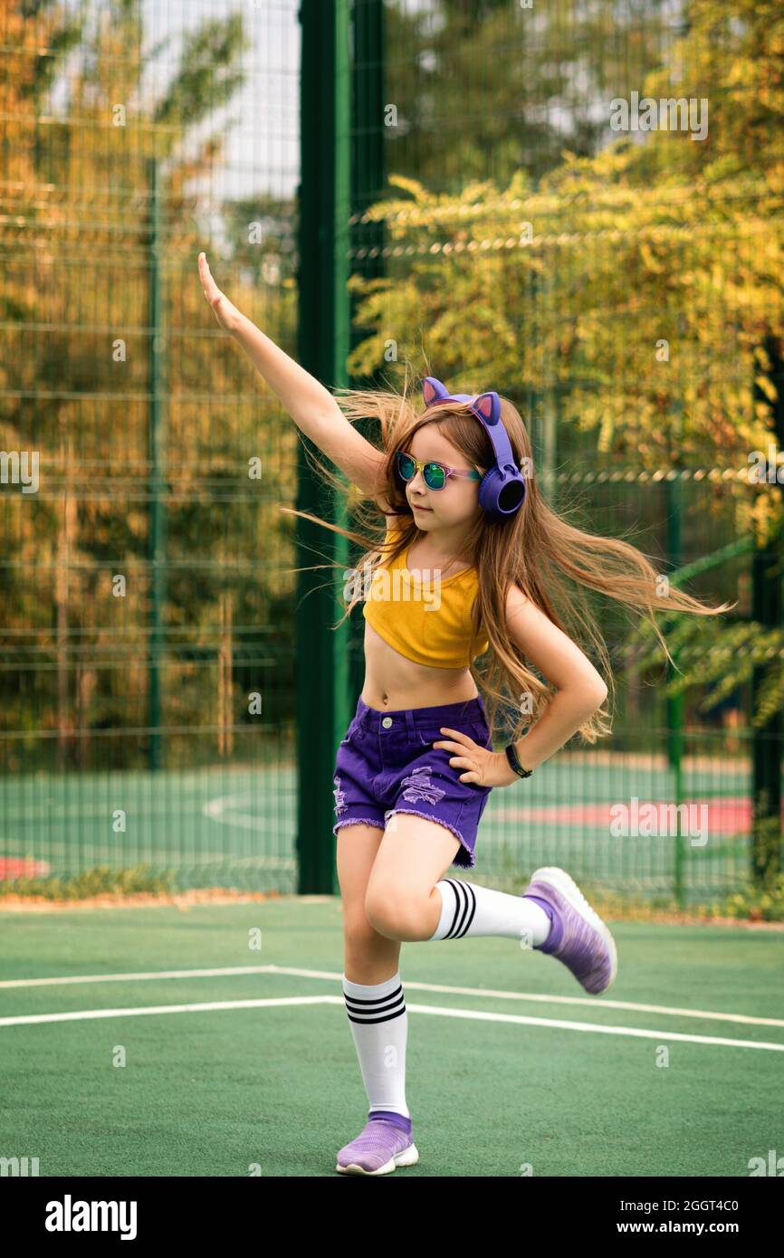 Girl wearing trendy fashionable sportswear and rainbow eyeglasses dancing on sports ground. Fashion pretty cool preteen schoolgirl with wireless headphones listening to music. Soft focus Stock Photo A...