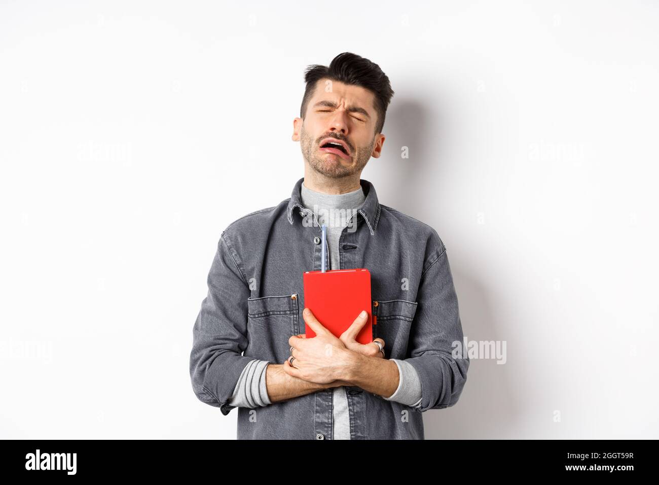 Sad cryig man holding red diary and sobbing, miserable guy carry journal with him, standing against white background Stock Photo