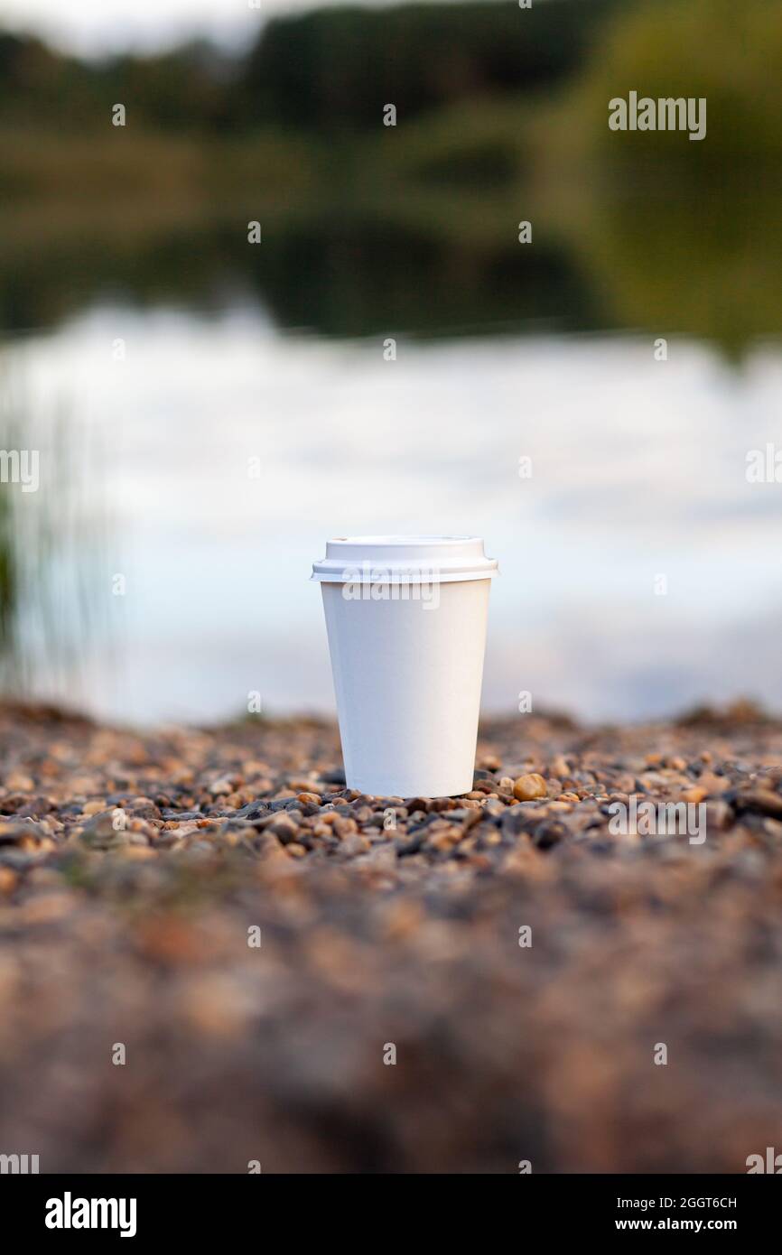 A white paper cup of coffee or tea stands on the rocky shore of the lake. A mug of hot drink on a pebble. Beautiful nature background of the lake in t Stock Photo