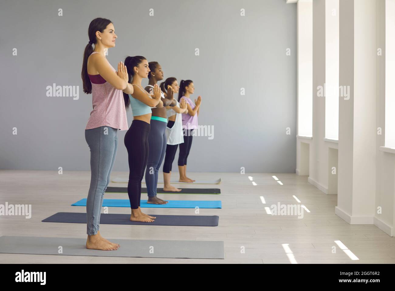 Group of young women standing on mats and doing Namaste during yoga class at gym Stock Photo